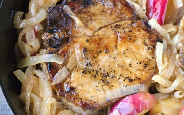 An Apple Pork Chop in a skillet with caramelized onions and apple slices