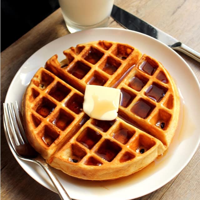 Greek Yogurt Waffle topped with syrup and a pad of butter on a white plate with a fork and butter knife. 