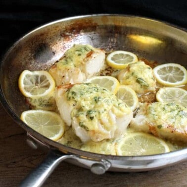Cod Fish covered in a lemon sauce and surrounded by lemon slices in a skillet.