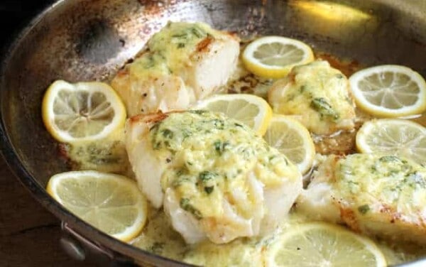 Cod Fish covered in a lemon sauce and surrounded by lemon slices in a skillet.