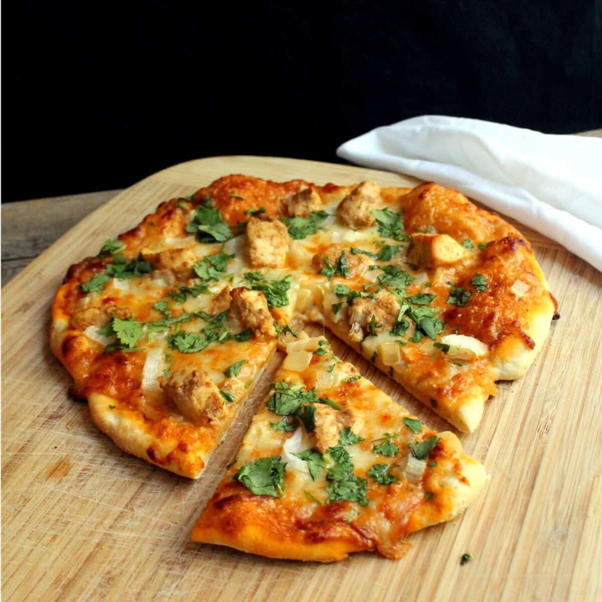 A chicken tikka masala pizza on a wooden cutting board with a slice cut out.