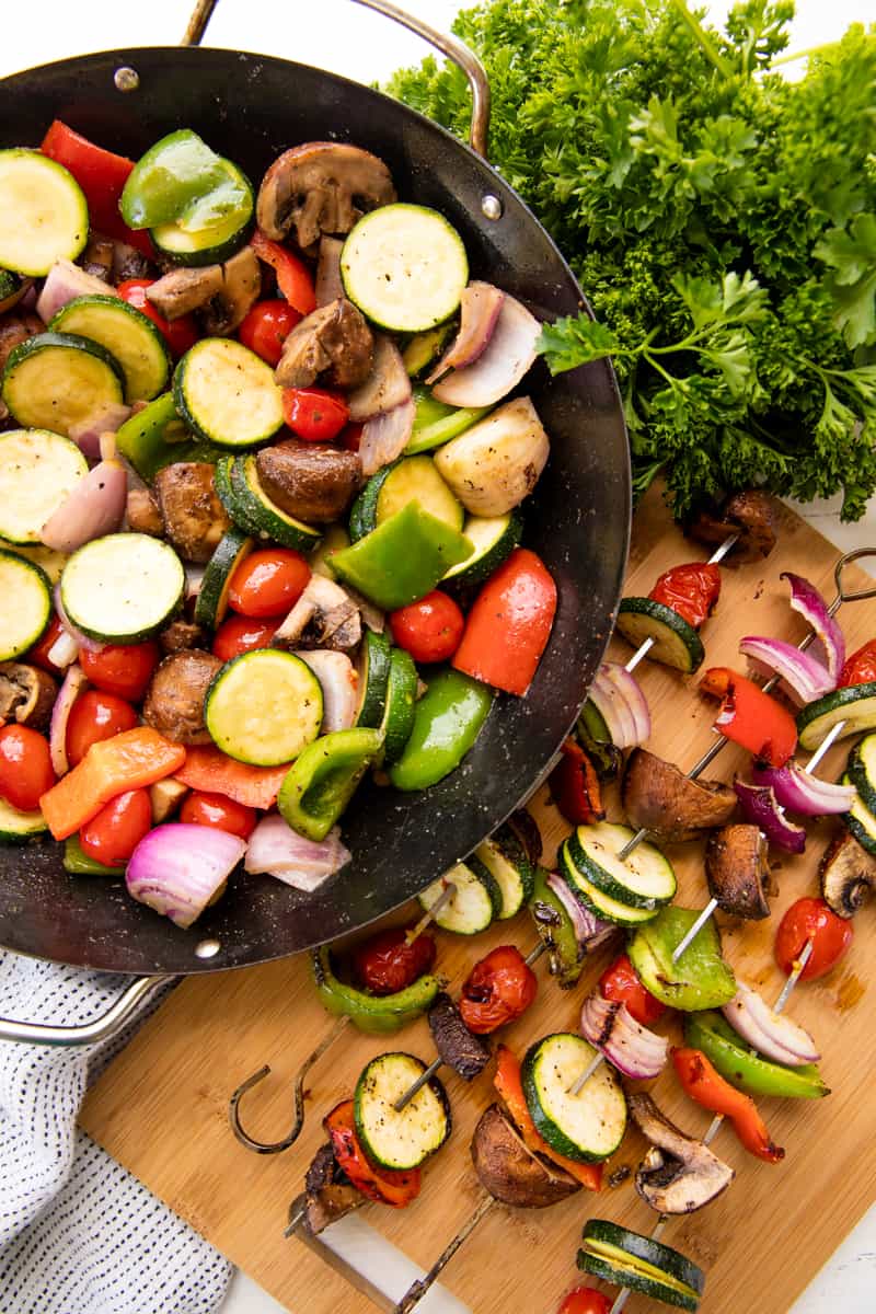 Fresh vegetables tossed with seasoning in a grill pan and grilled veggie kebabs on metal skewers on a cutting board