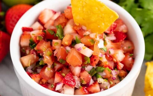 A tortilla chip is dipped into a bowl of strawberry salsa, surrounded by fresh strawberries, lemon, cilantro and tortilla chips