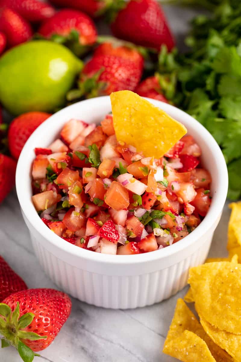 A tortilla chip is dipped into a bowl of strawberry salsa, surrounded by fresh strawberries, lemon, cilantro and tortilla chips