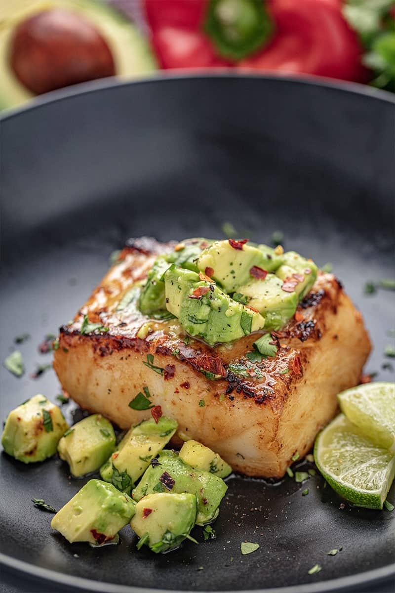 A closeup of Avocado Salsa Sea Bass in a skillet with avocado salsa and slices of lime