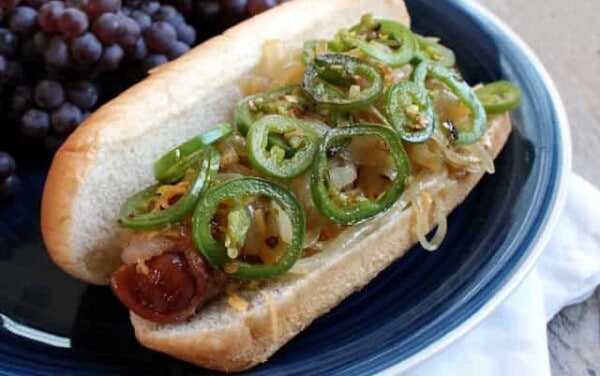 Bacon-wrapped hot dog in a hot dog bun covered in onions and jalapeno slices.