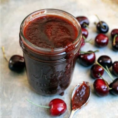 Cherry BBQ Sauce in a mason jar on a counter with a spoon full of sauce and cherries.