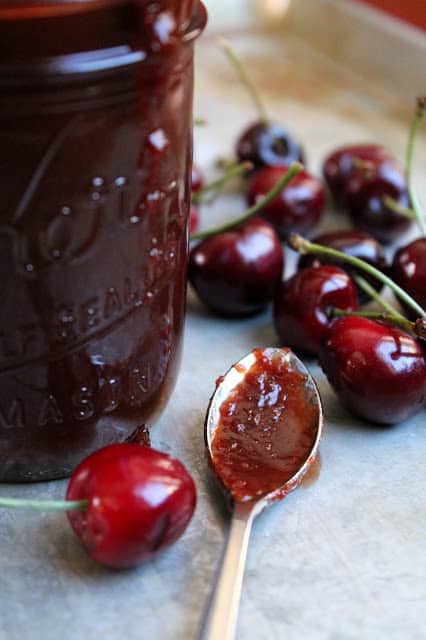 A jar of cherry bbq sauce sitting next to cherries and a spoon full of the sauce.