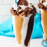 Chocolate Peanut Butter MilkShake in a tall glass with a straw topped with crumbles up Reese's peanut butter cups, whipped cream, and full Reese's peanut butter cup on top.