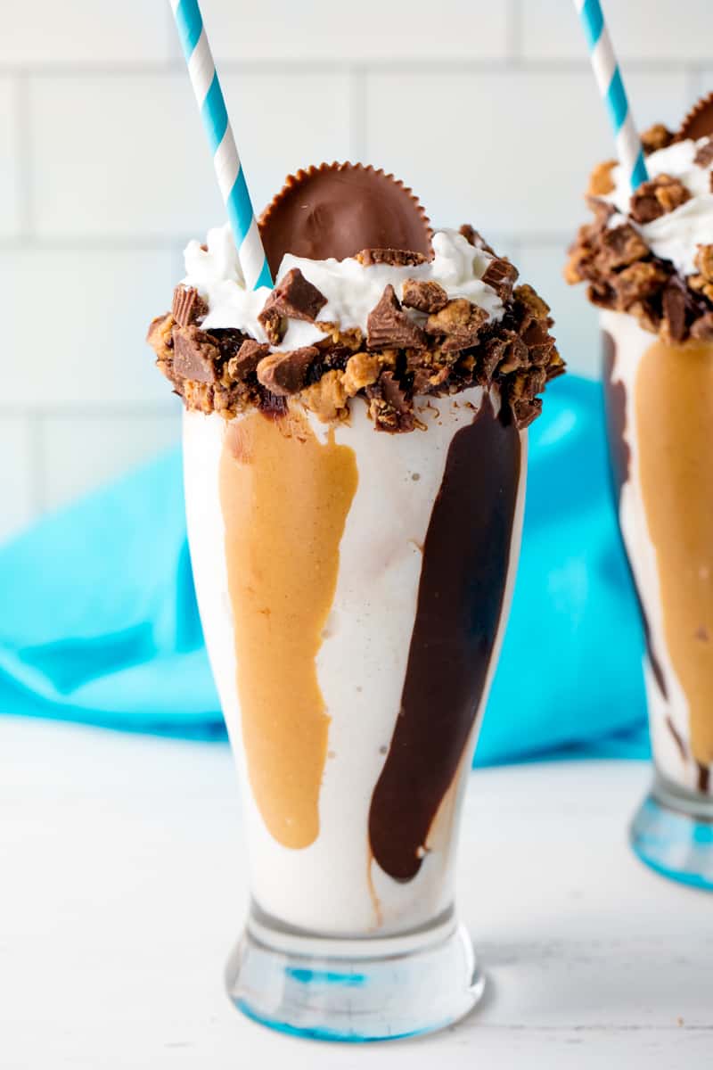 Chocolate Peanut Butter MilkShake in a tall glass with a straw topped with crumbles up Reese's peanut butter cups, whipped cream, and full Reese's peanut butter cup on top.