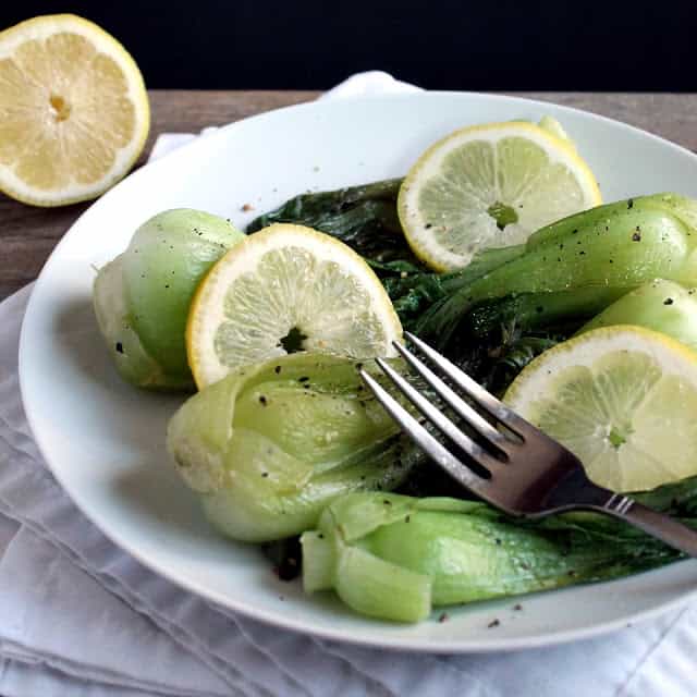 Cooked bok choy on a white plate with lemon slices on it.