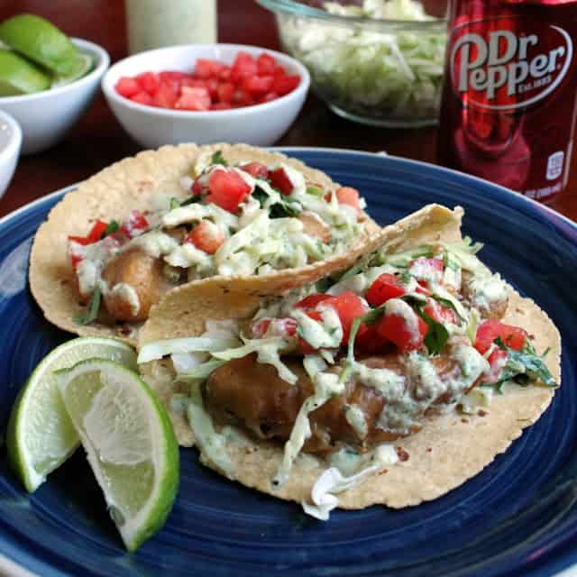 Dr Pepper Battered Fish Tacos on a blue plate by two lime wedges.