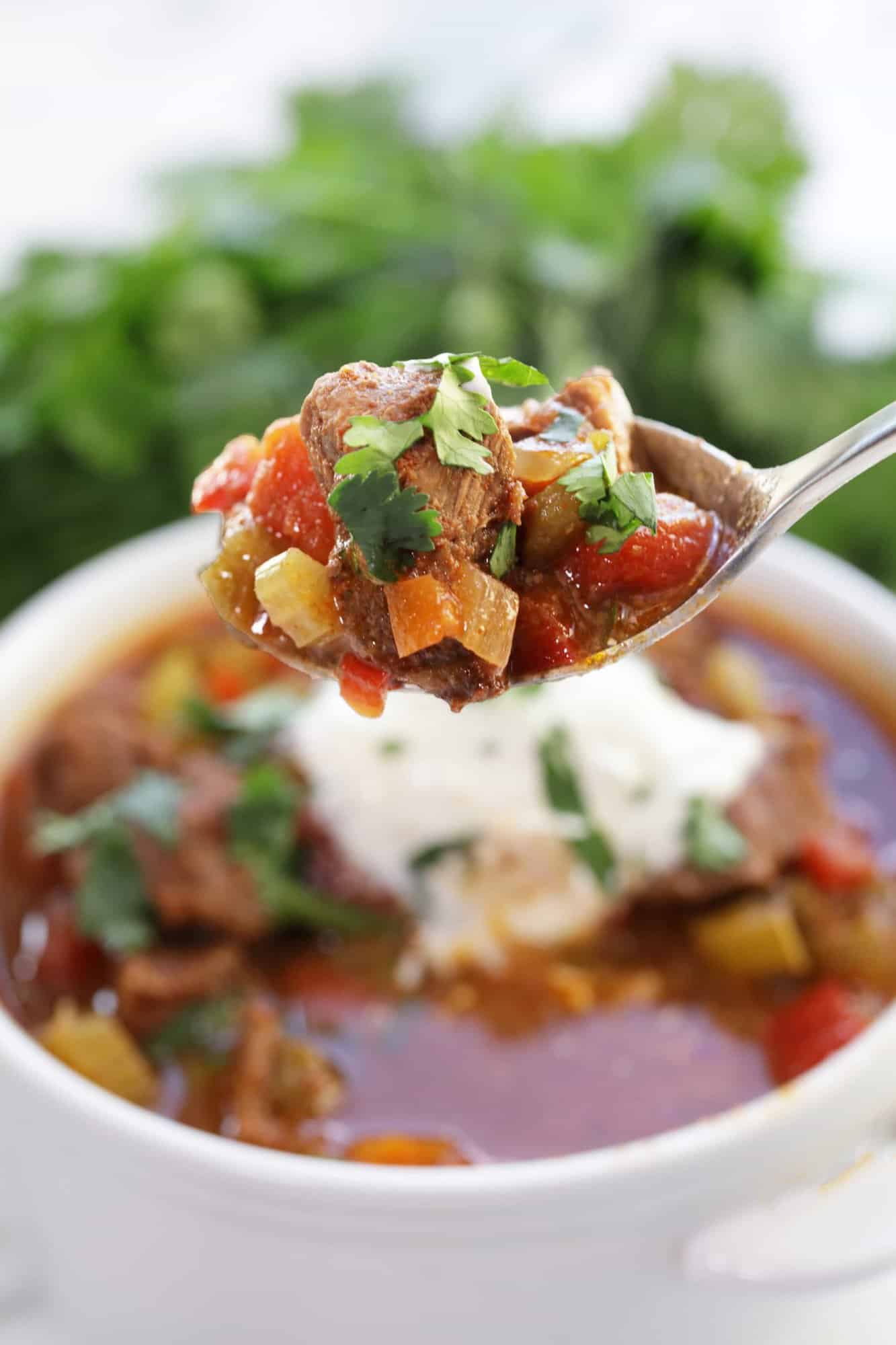 A spoon lifts a bite of Slow Cooker Texas Beef Chili with chunks of beef, slow cooked tomatoes, bell pepper, carrots onion and celery and topped with fresh cilantro.