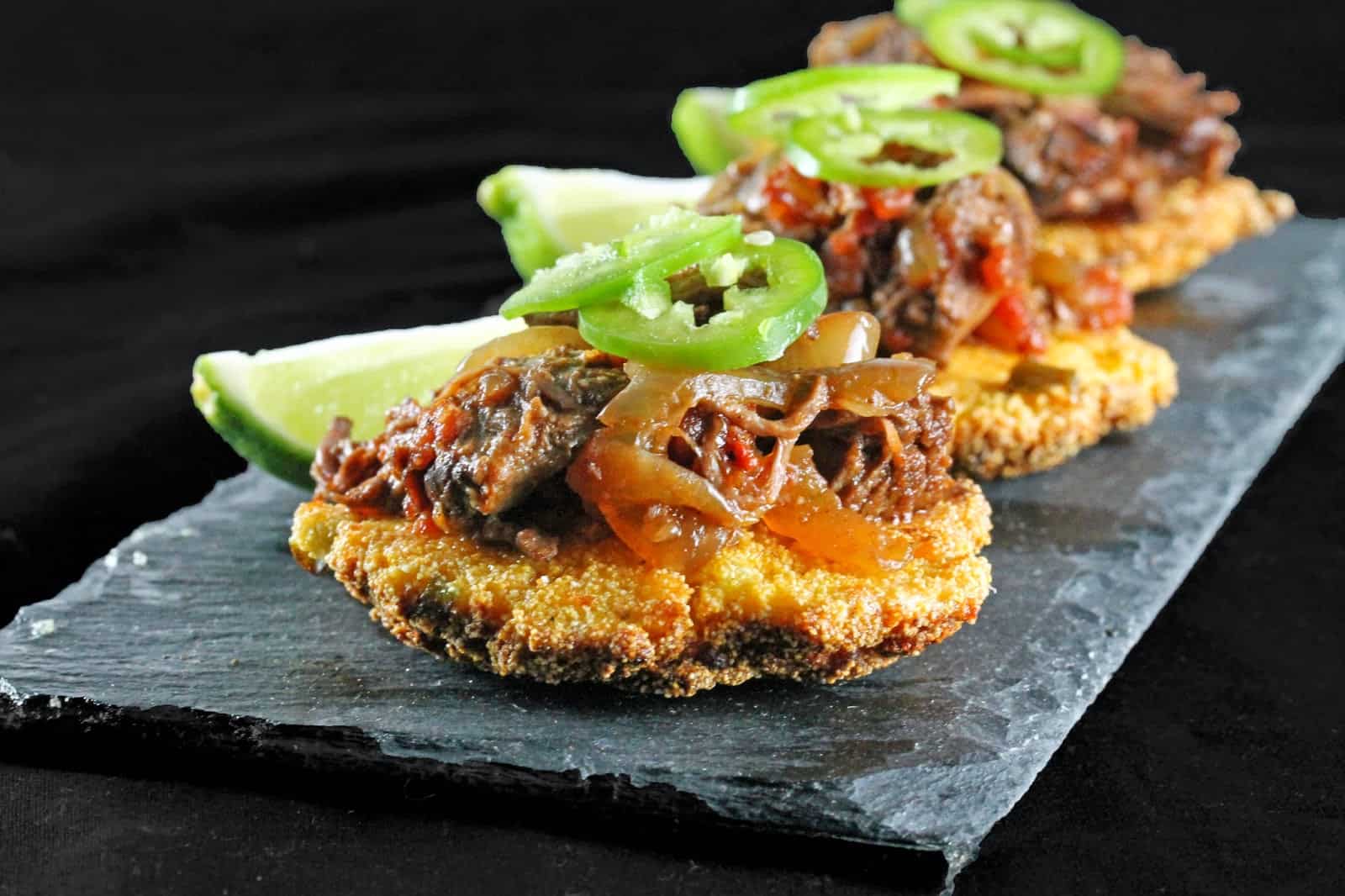Slow Cooker Ancho Chili Shredded Beef over Corn Fritters is garnished with sliced fresh jalapenos and limes