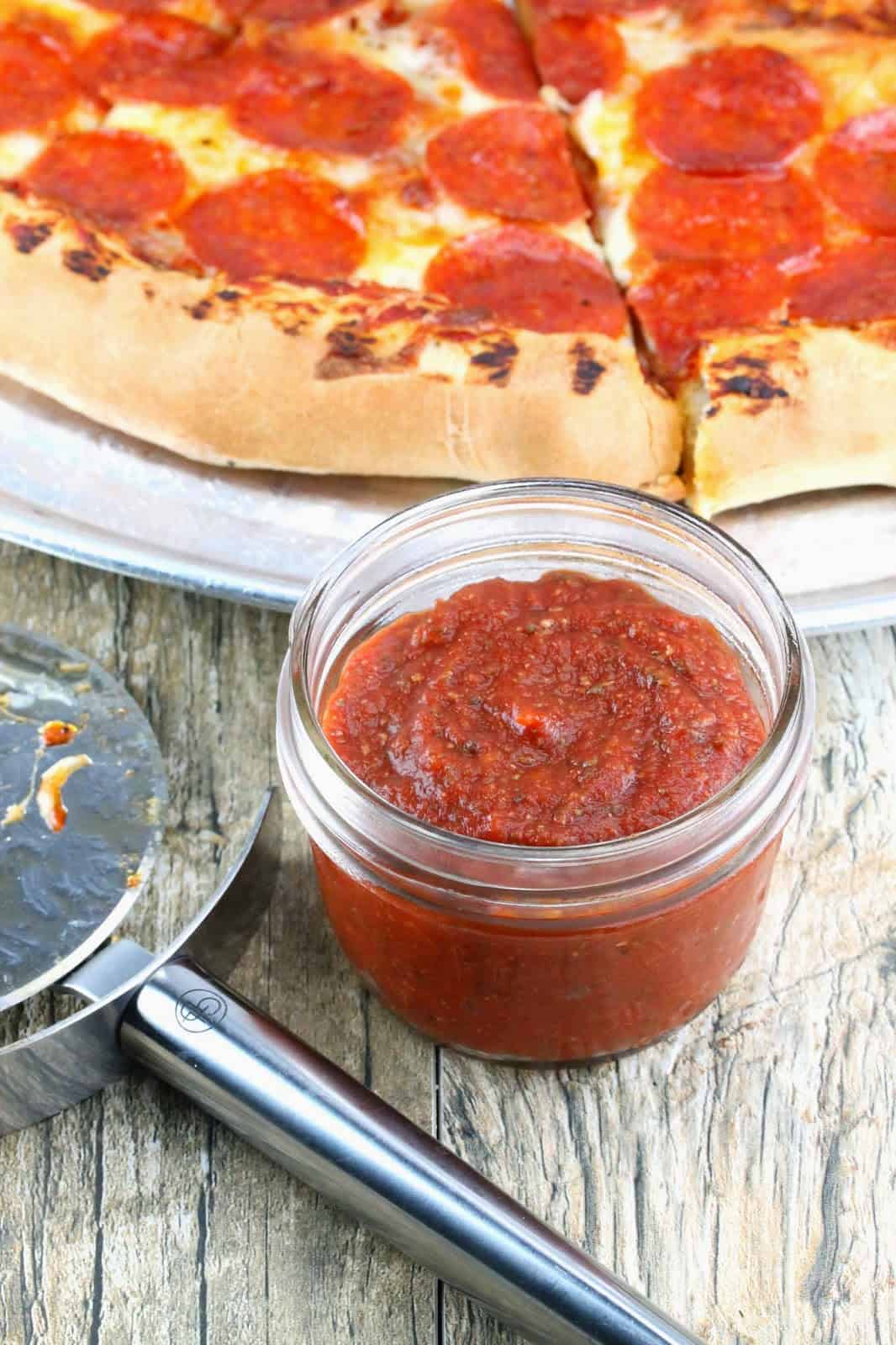 A small mason jar full of pizza sauce next to a pizza cutter and a pizza.