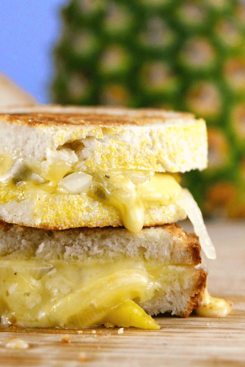 A close up view of a 
grilled cheese sandwich that has been cut in half and stalked on top of each other. It has caramelized pineapple, onions, and jalapeno. There is a pineapple in the background. 