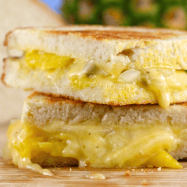 A close up view of a grilled cheese sandwich that has been cut in half and stalked on top of each other. It has caramelized pineapple, onions, and jalapeno.