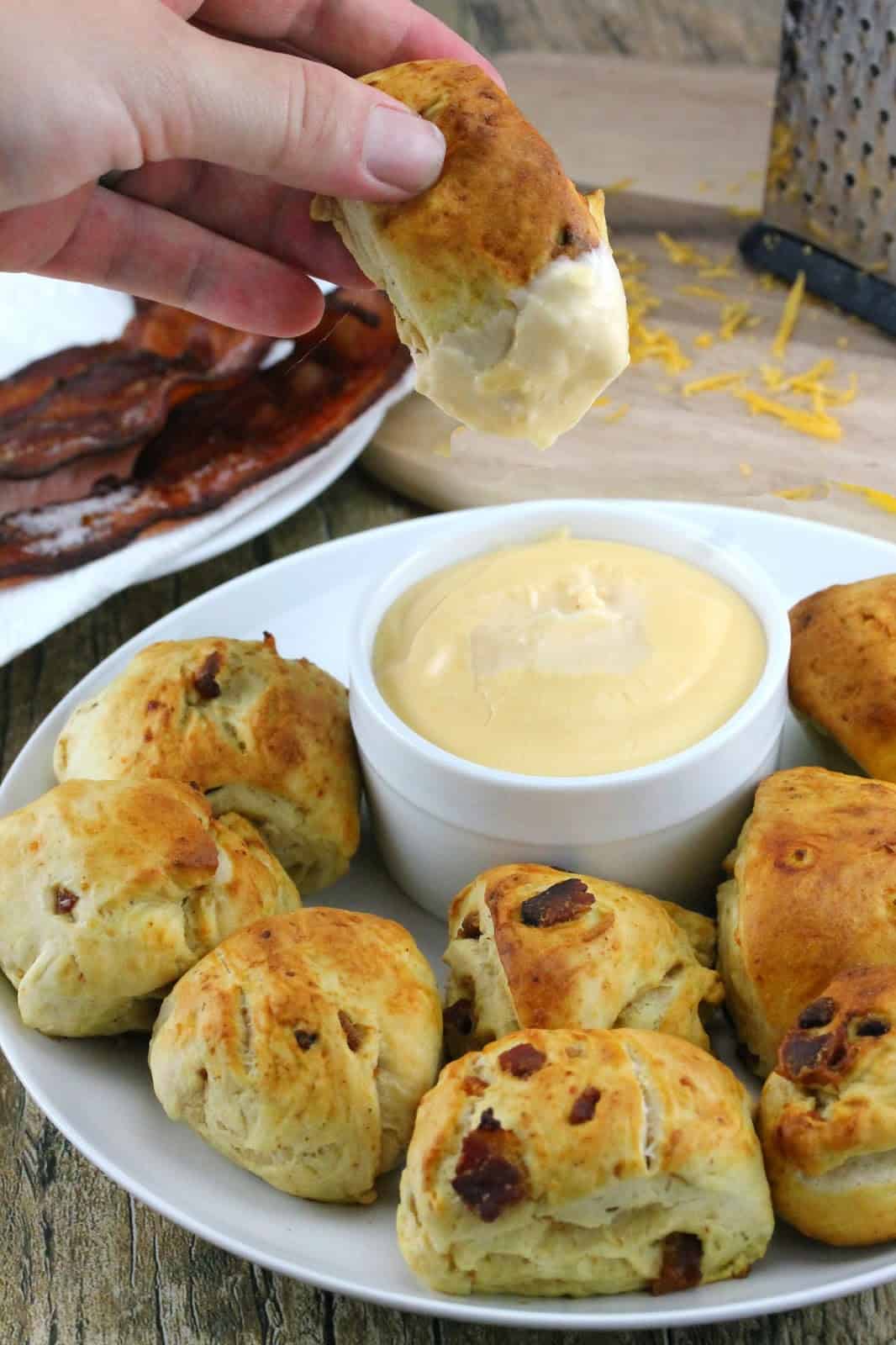 Bacon cheddar pretzel bites on a white plate with a dipping sauce. One bacon cheddar pretzel bite is being pulled out of the dip.