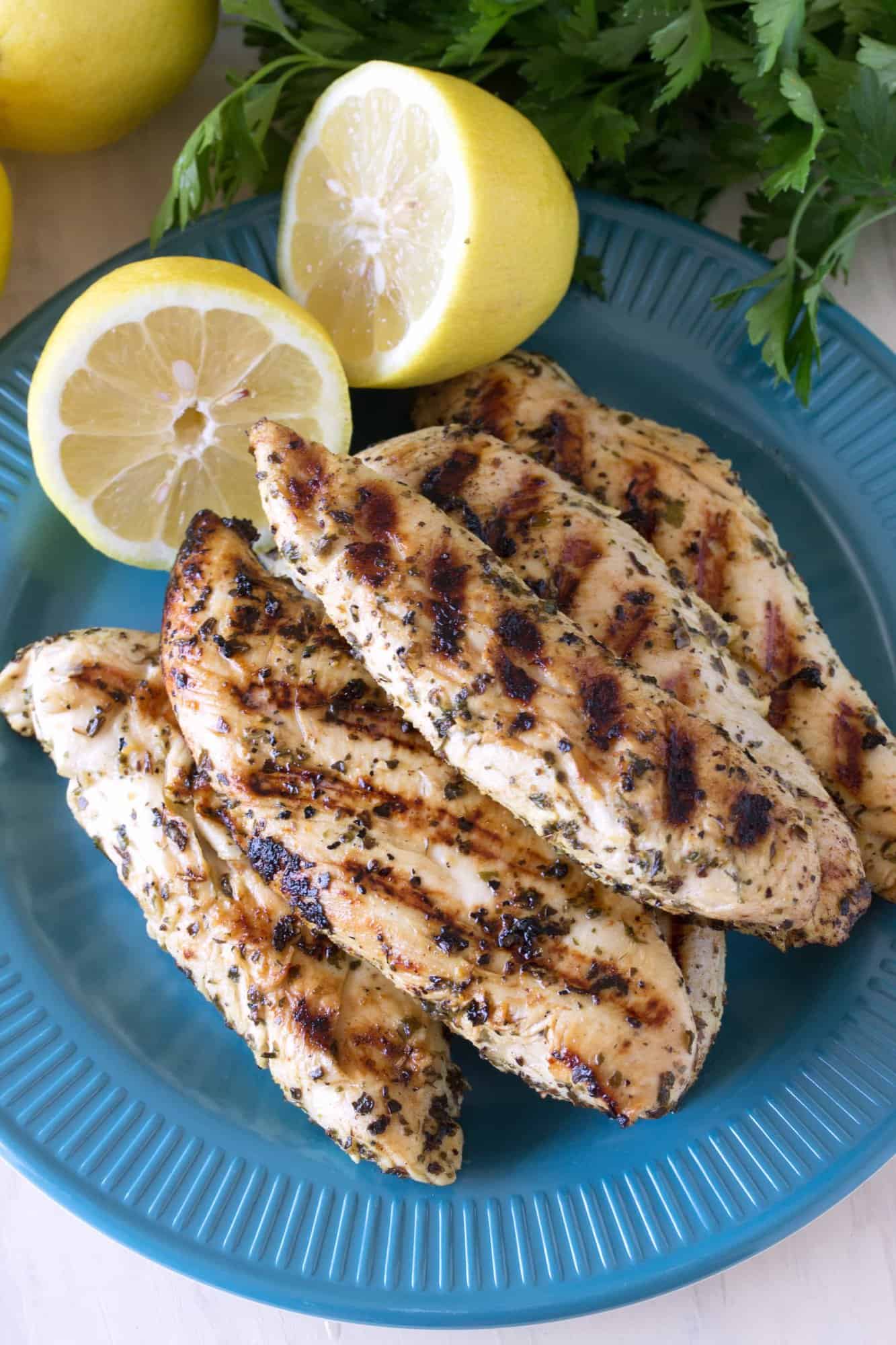 Grilled chicken tenders stacked on a blue plate with two half lemons on it..