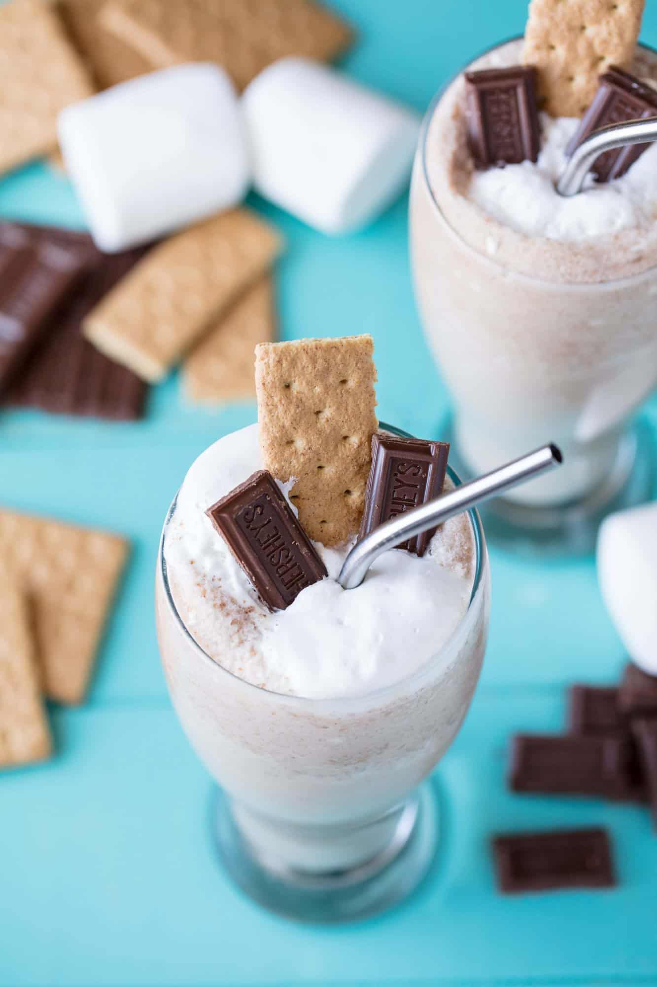 S'mores Frappes in a glass with a metal straw, topped with marshmallow cream, pieces of Hershey's bar and graham cracker