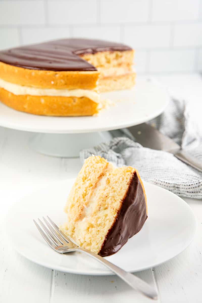 Slice of boston cream pie on a white plate with a fork on it. 