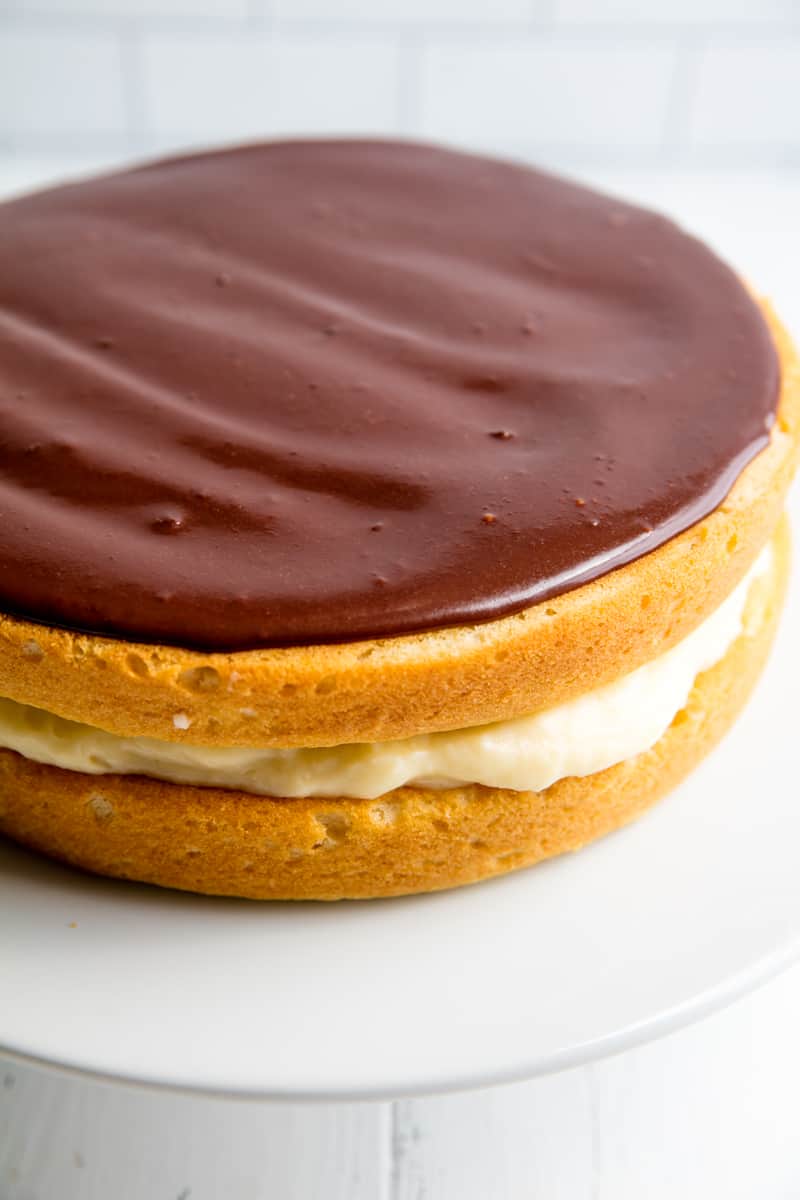 Boston Cream Pie with a chocolate ganache on top sitting on a cake stand. 