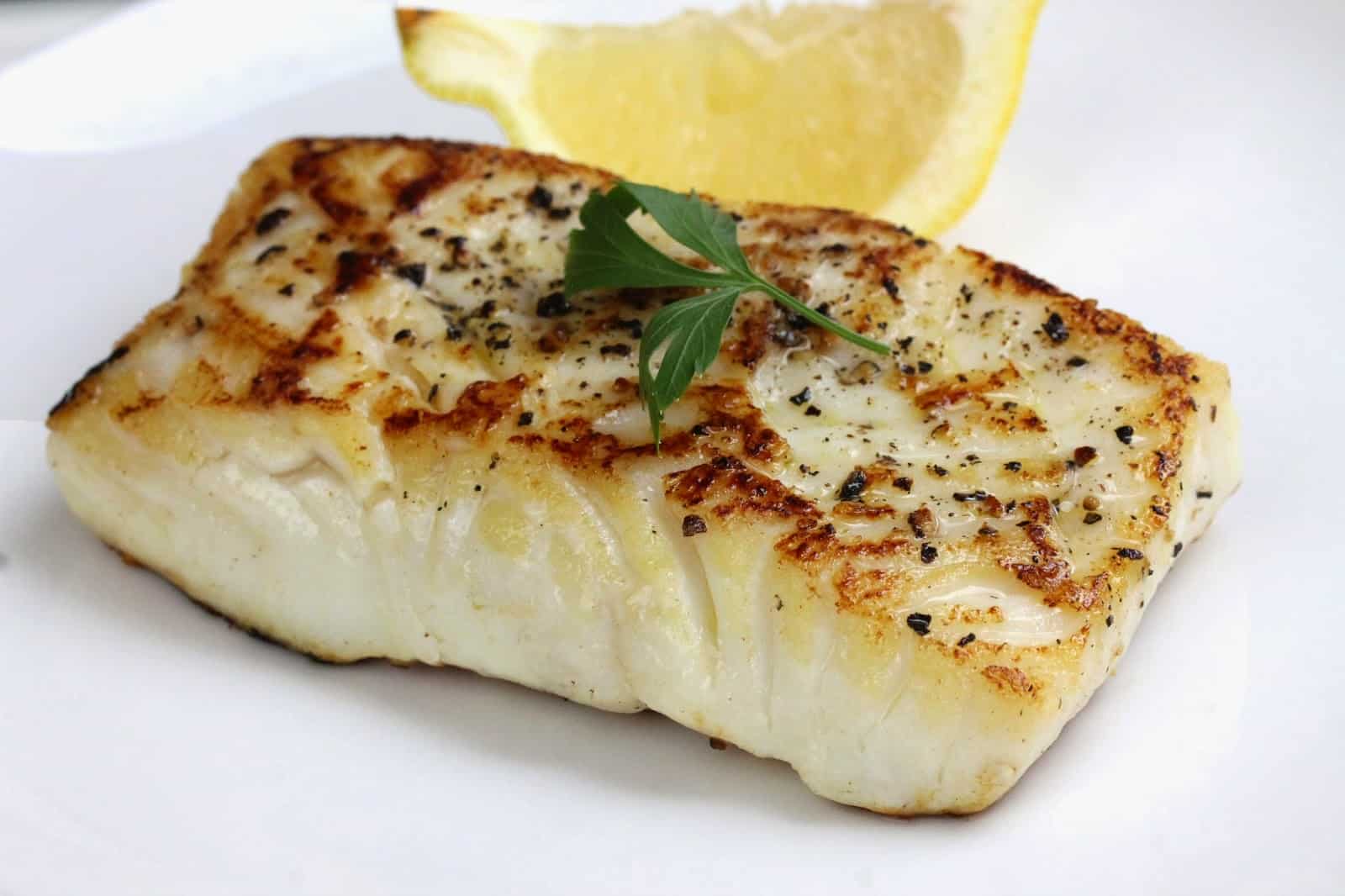 Pan seared fish on a white plate with a lemon wedge by it.