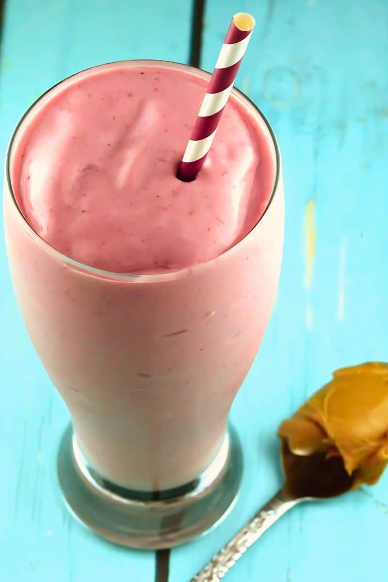 A pink peanut butter and jelly smoothie with a spoon of peanut butter laying next to it.