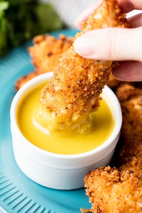 Cajun fried chicken strip being dipped into a container of honey mustard on a blue plate