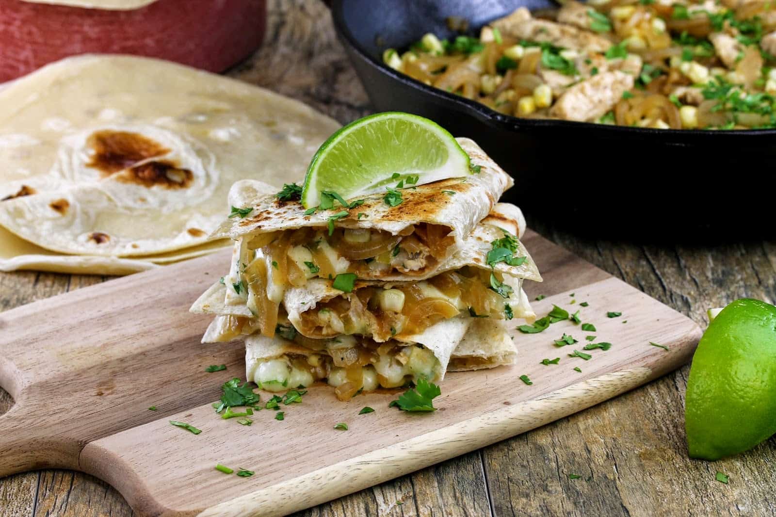 A stack of chicken-lime quesadillas spilling over with cheese, onions, corn and chicken and garnished with fresh cilantro and a lime wedge all on a cutting board.