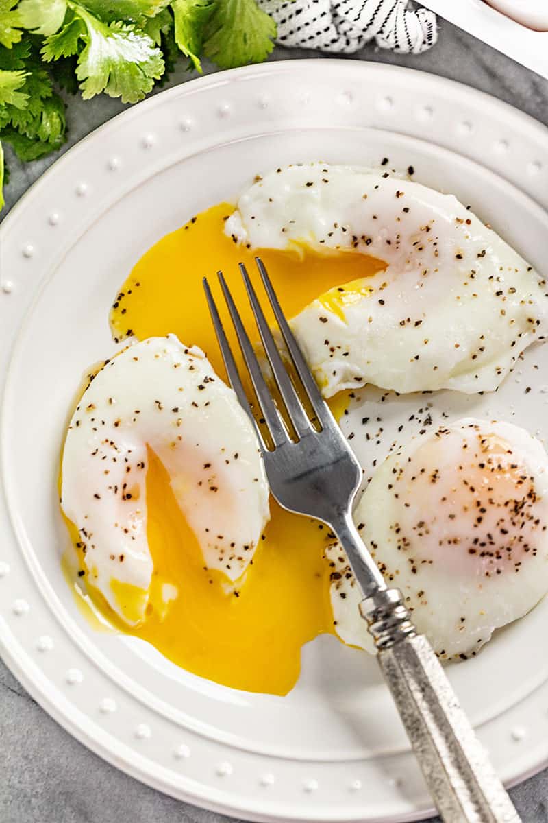 A plate of poached eggs seasoned with salt and pepper. Yolks are cut into with a fork and runny