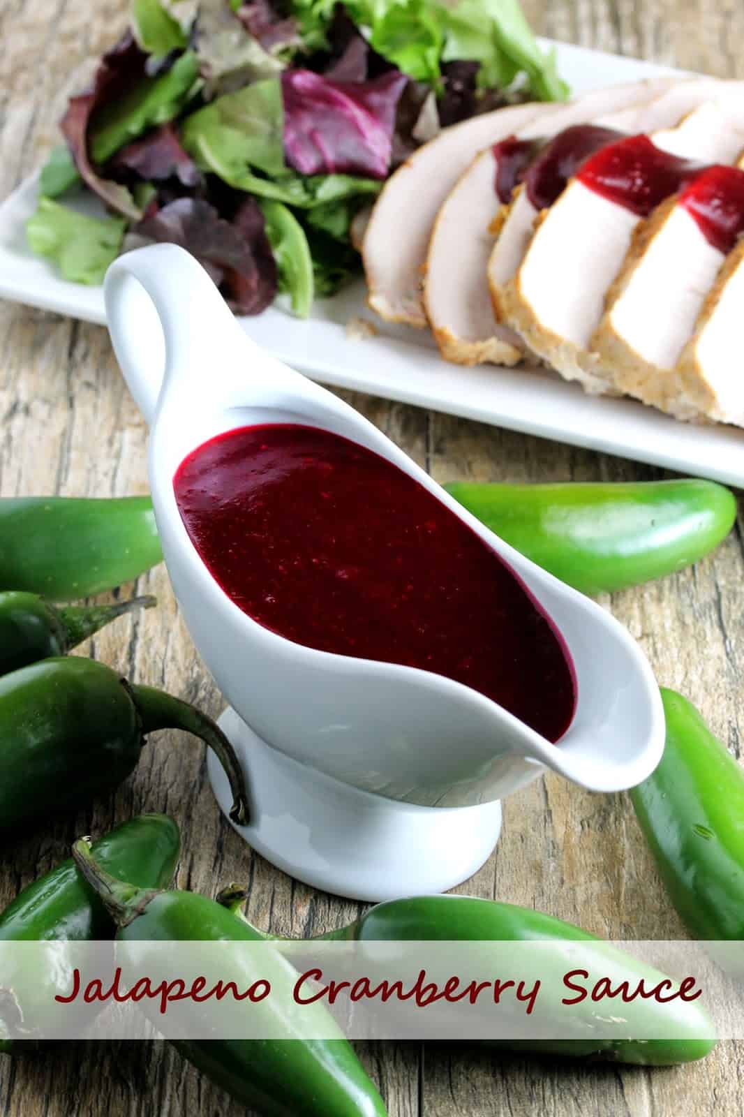 Jalapeno Cranberry Sauce in the background is drizzled over turkey breast on a platter with salad greens. A gravy boat full of Jalapeno Cranberry Sauce in the foreground surrounded by fresh jalapenos.