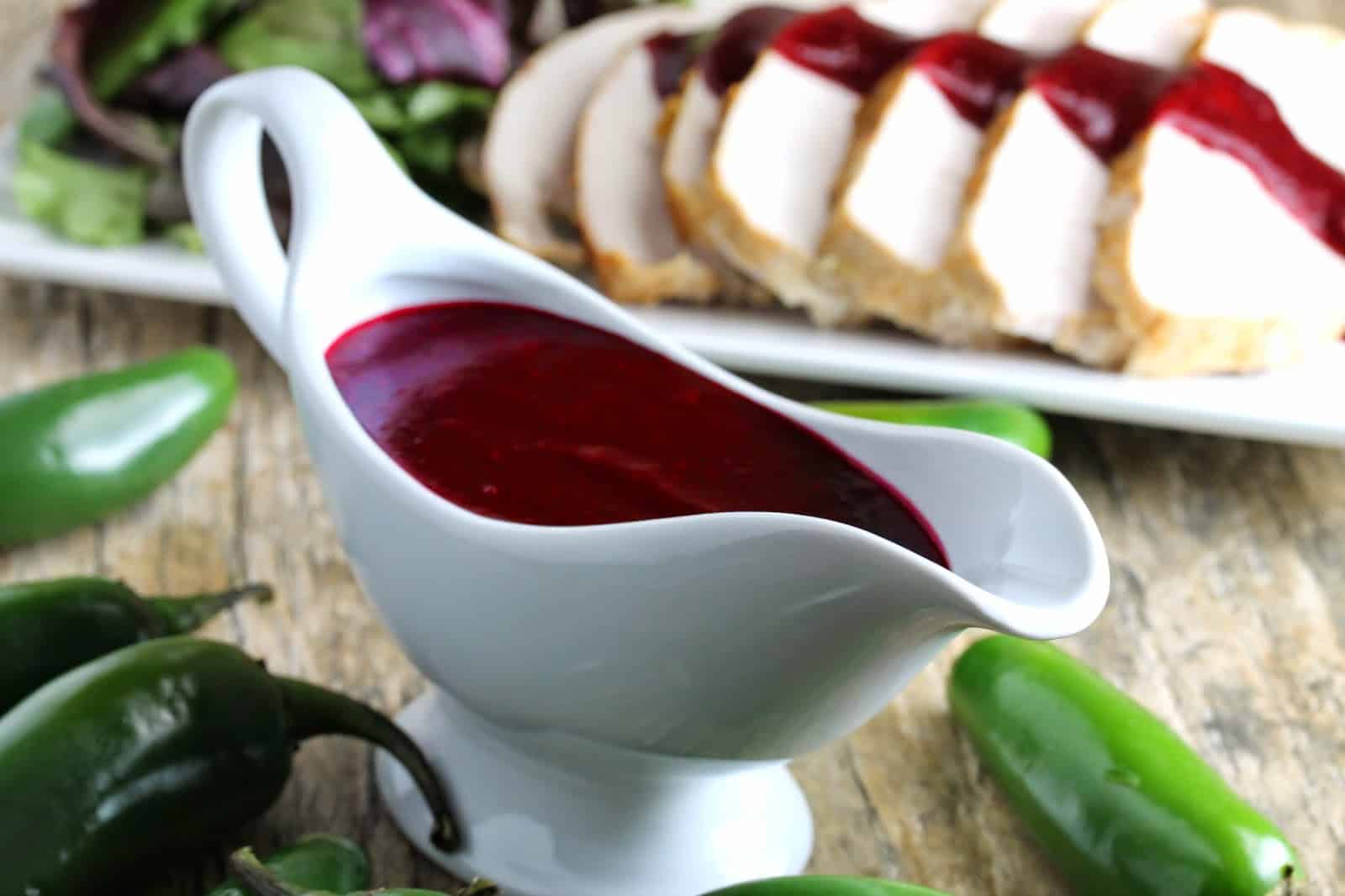 Jalapeno Cranberry Sauce in the background is drizzled over turkey breast on a platter with salad greens. A gravy boat full of Jalapeno Cranberry Sauce in the foreground surrounded by fresh jalapenos.