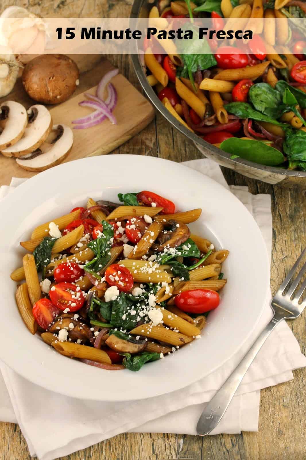 Pasta made with tomatoes, spinach, onions and mushrooms, and topped with crumbled feta cheese in a white bowl next to a cutting board with toppings on it and a metal bowl full of the pasta. 