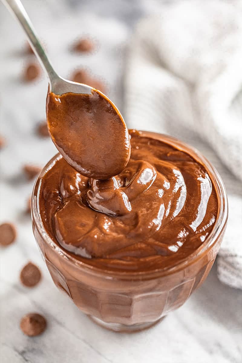 Bird's eye view of a spoon drizzling Chocolate Pudding in a glass dish full of chocolate pudding.