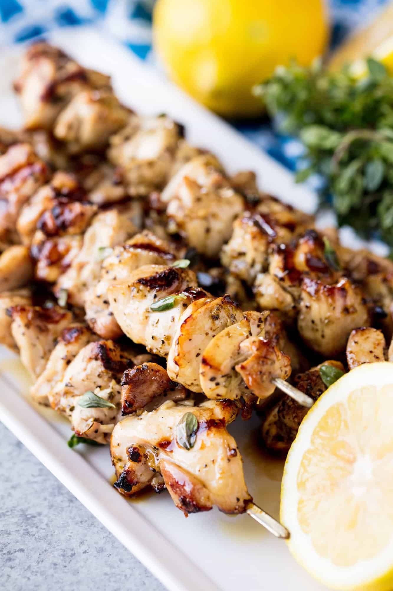 Grilled Lemon Herb Chicken Thigh Skewers off the grill, stacked on a serving platter with a lemon slice with fresh lemon and oregano nearby