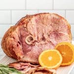 Cooked ham on a serving plate with sliced oranges and rosemary.
