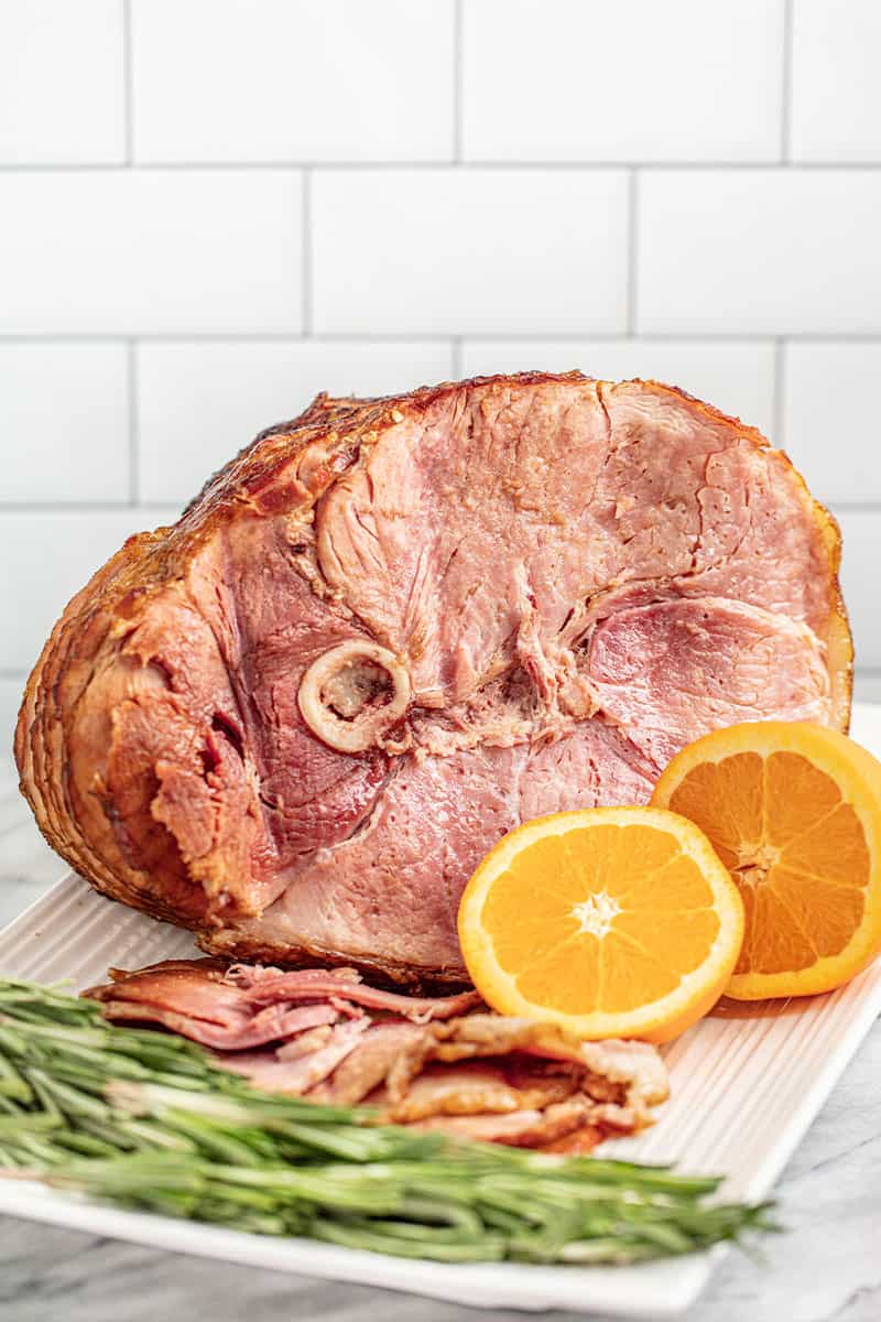 Cooked ham on a serving plate with sliced oranges and rosemary. 