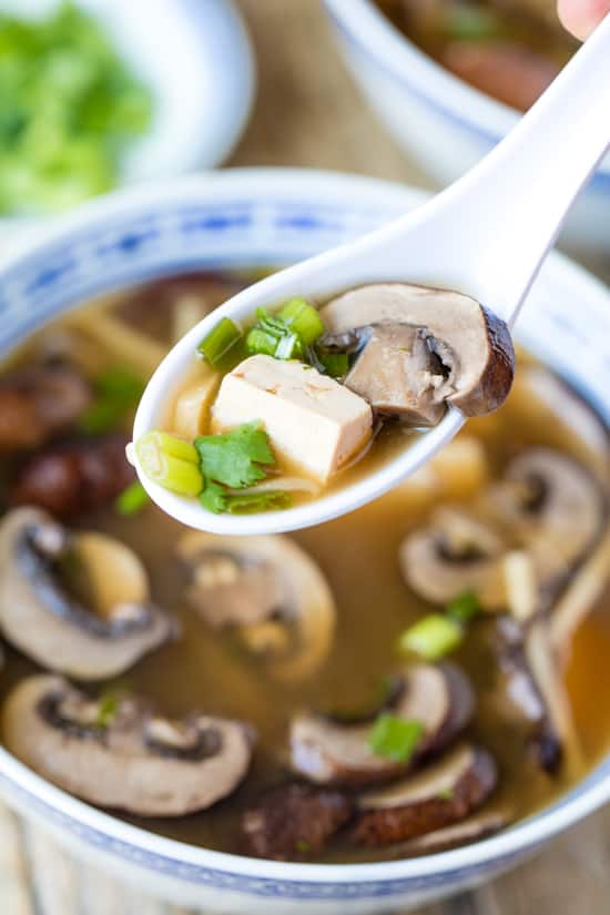 A soup spoon with a bite of Slow Cooker Chinese Hot and Sour Soup is lifted from a bowl. Spoon is filled with mushroom, tofu, green onion, cilantro and bamboo shoot