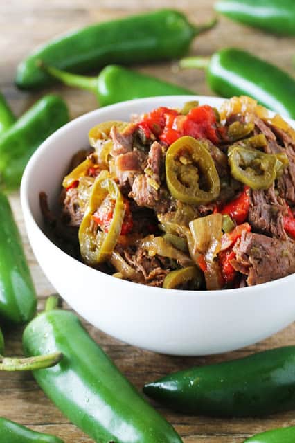 Slow cooked jalapeno beef in a white bowl surrounded by jalapeno peppers. 