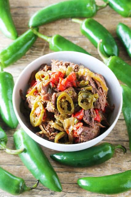 Bird's eye view of Slow cooked jalapeno beef in a white bowl surrounded by jalapeno peppers. 