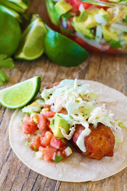 An above view of Crispy battered cod is topped with avocado salsa, simple slaw and jalapeno, and on a corn tortilla with a lime wedge sitting next to it.