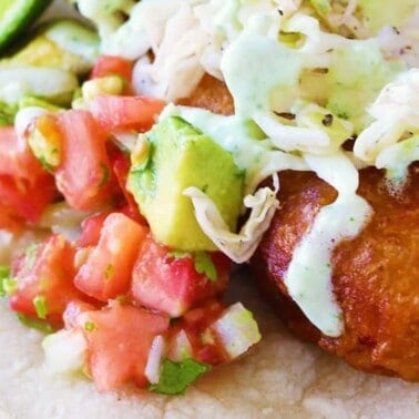 Close-up view of Battered Cod Fish Taco garnished with avocado, fresh salsa, and jalapeño crema on a corn tortilla.