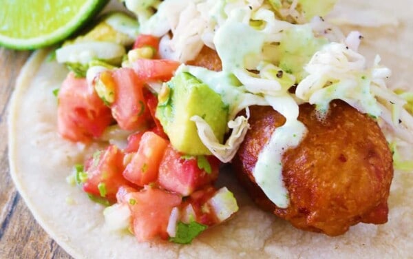 Close-up view of Battered Cod Fish Taco garnished with avocado, fresh salsa, and jalapeño crema on a corn tortilla.