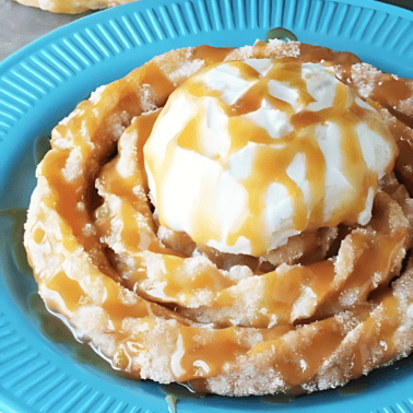 A close up view of a churro funnel cake on a blue plate, topped with whipped cream and drizzled with caramel sauce.