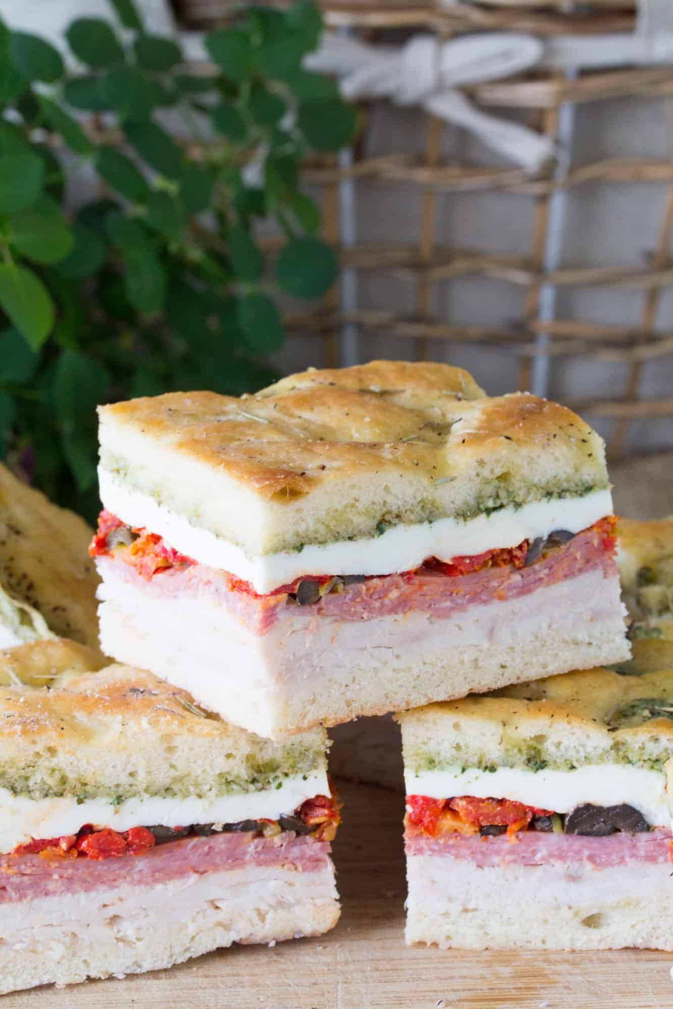 A stack of three Pressed Italian Sandwichs made of sliced chicken breast, sliced salami, sundried tomatoes, olives, pepperocinis, red bell peppers, and fresh mozzarella with a pesto sauce\.
