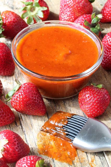 Sweet and smokey barbecue sauce served up in a glass container surrounded by strawberries. 