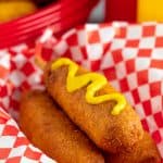 Three Corn Dogs in a basket with the top one topped with mustard.