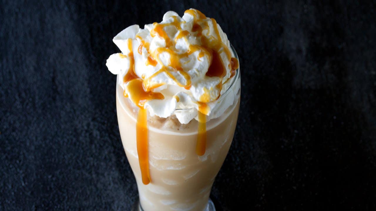Angled view of Salted Caramel Frappes in a curved clear glass topped with whipped cream and drizzled with caramel syrup.