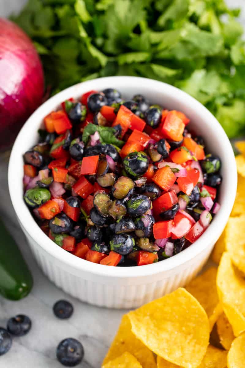 Blueberry Bell Pepper Salsa in a white bowl, surrounded by fresh cilantro, onion, jalapeno, blueberries and tortilla chips
